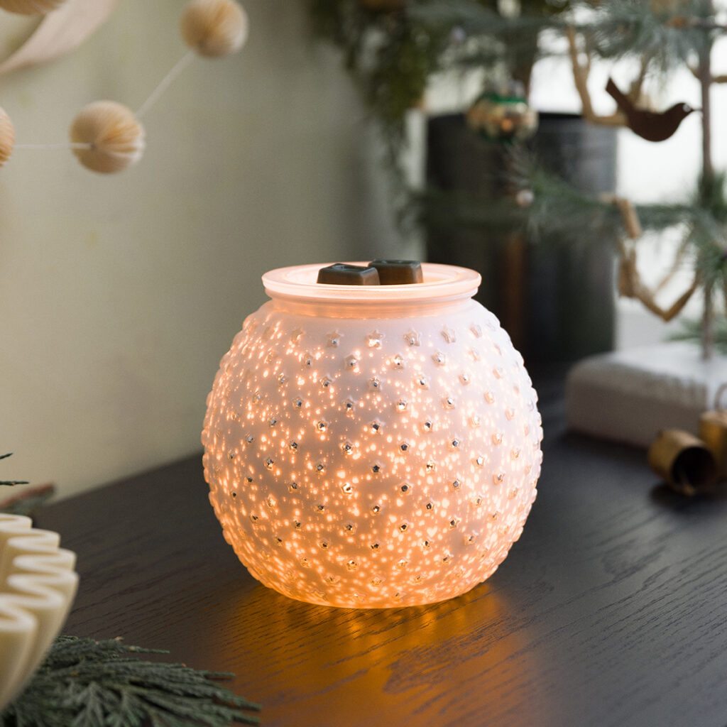 The Snowdrop Warmer by Scentsy glowing warmly with a golden light shining through its star-dotted hobnail glass. Creates the coziest holiday glow — perfect for winter decorating, relaxing nights, and filling your home with your favorite Scentsy scent.”