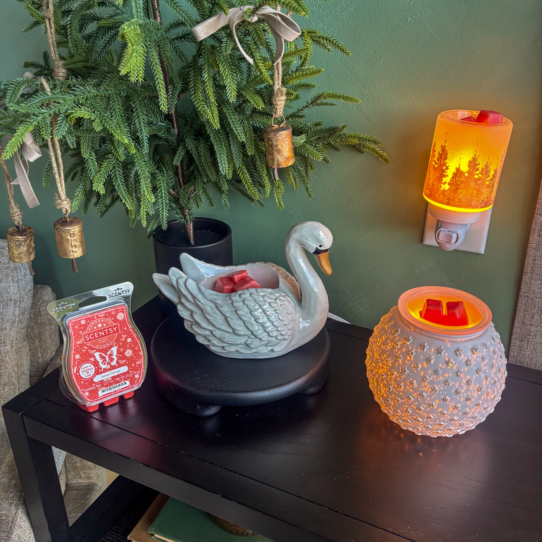 Cozy little holiday Scentsy moment — my All Is Calm Mini Warmer glowing next to my swan and hobnail warmers, all melting red holiday wax bars. Totally giving cozy cabin vibes with the mini tree and rustic bells — perfect warm winter home setup.
