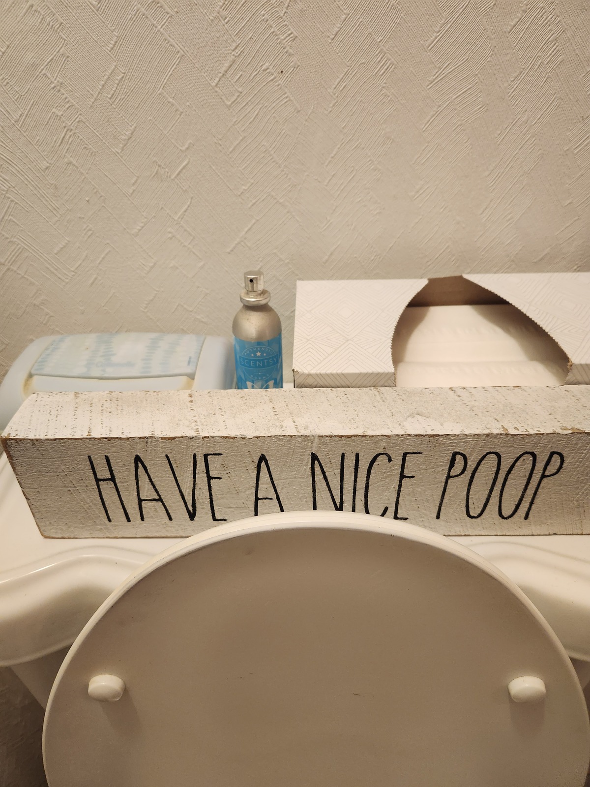 Scentsy Room Spray on the back of a toilet beside tissues and a funny bathroom sign that says ‘Have a Nice Poop.’ Perfect for quick bathroom odor control and a touch of humor in home decor.