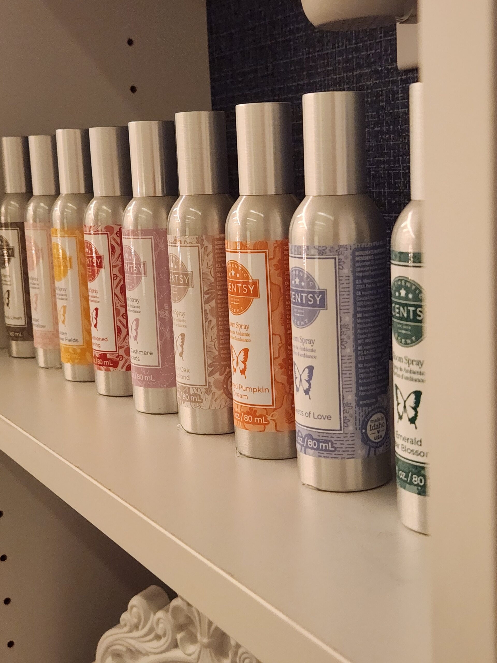 Line of colorful Scentsy Room Sprays displayed on a white shelf. Each aluminum bottle features a different fragrance label, showing the variety of scents available for instant home fragrance.