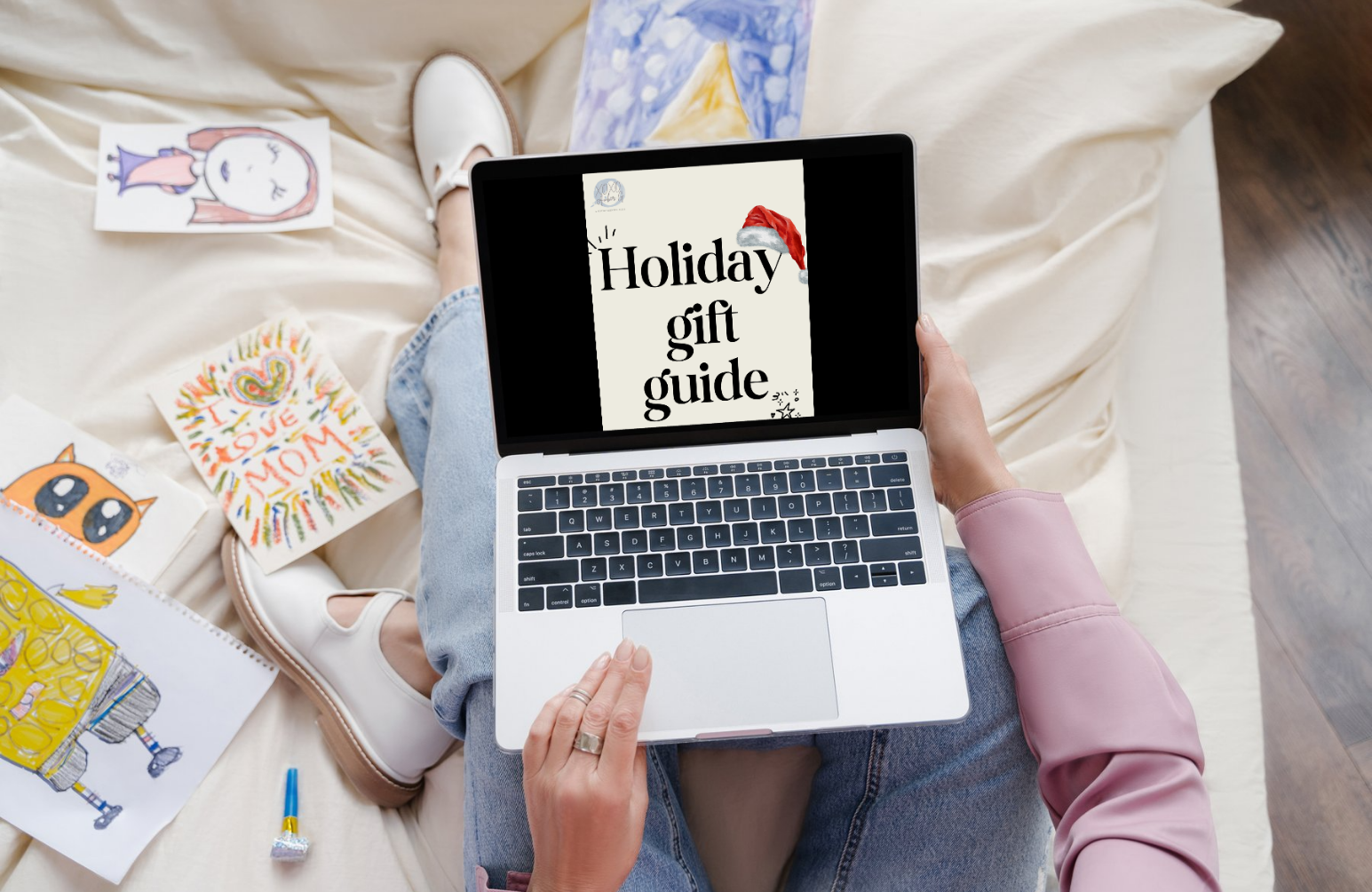 A woman sitting on a cozy bed with kids’ drawings around her, holding a laptop displaying Amber G’s “Holiday Gift Guide” cover with a Santa hat graphic. Warm, relaxed vibe that invites viewers to download the free holiday gift guide