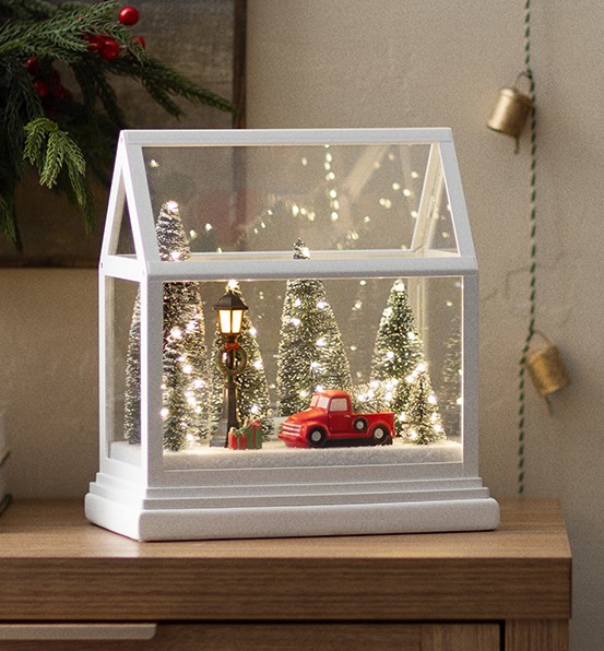 Close-up of the Dashing Through the Pines Limited Edition Holiday Warmer by Scentsy featuring a red truck, lamppost, presents, and snowy pine trees lit with sparkling white lights inside a glass house design.