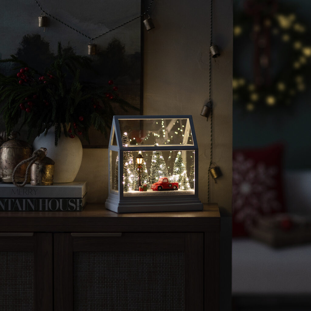 The Dashing Through the Pines Holiday Warmer by Scentsy glowing at night with twinkling lights around snowy trees and a red truck under a glass house design — the perfect cozy glow for winter evenings.