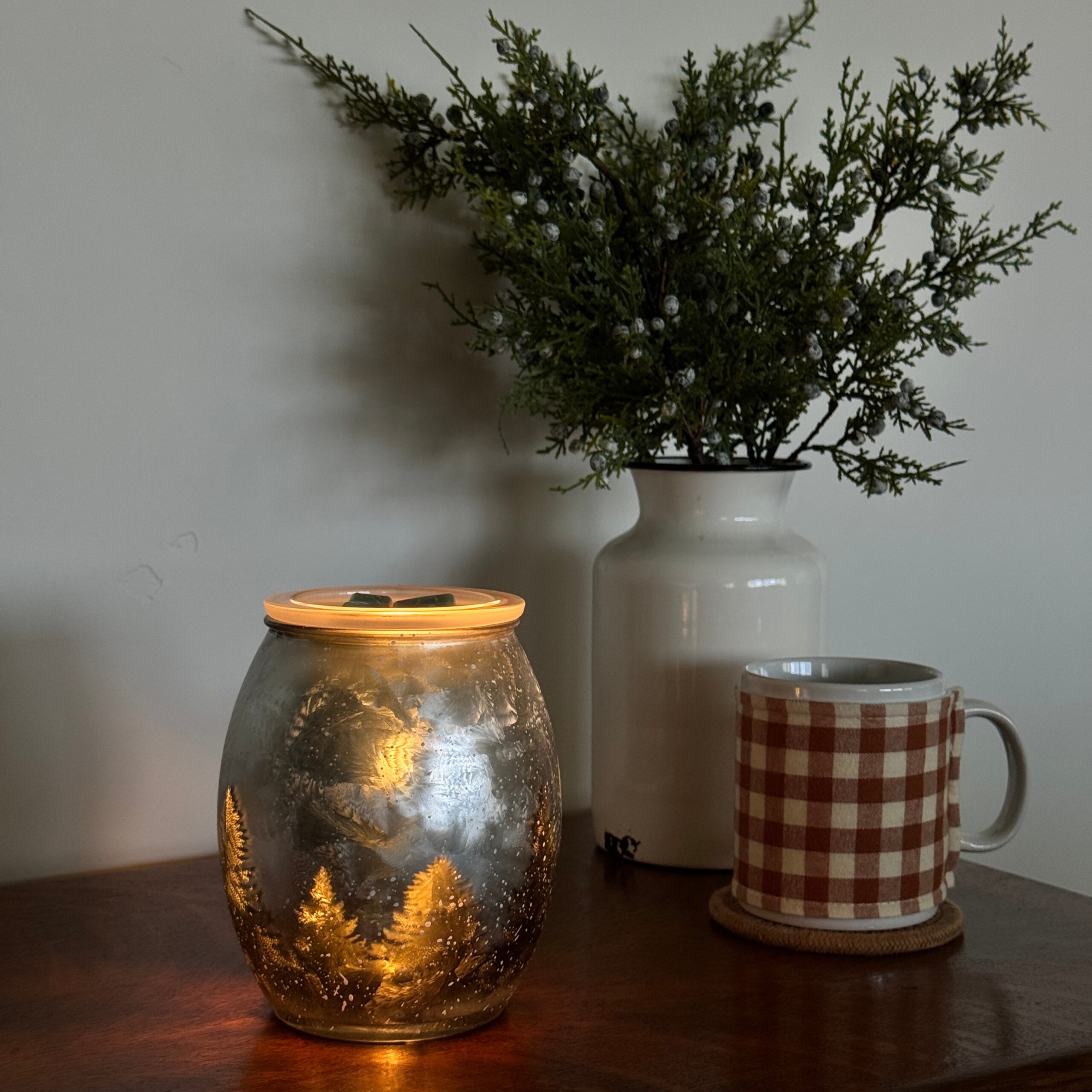 Lit Scentsy Wintercrest Warmer on a table next to a plaid mug and greenery, creating a cozy winter scene.