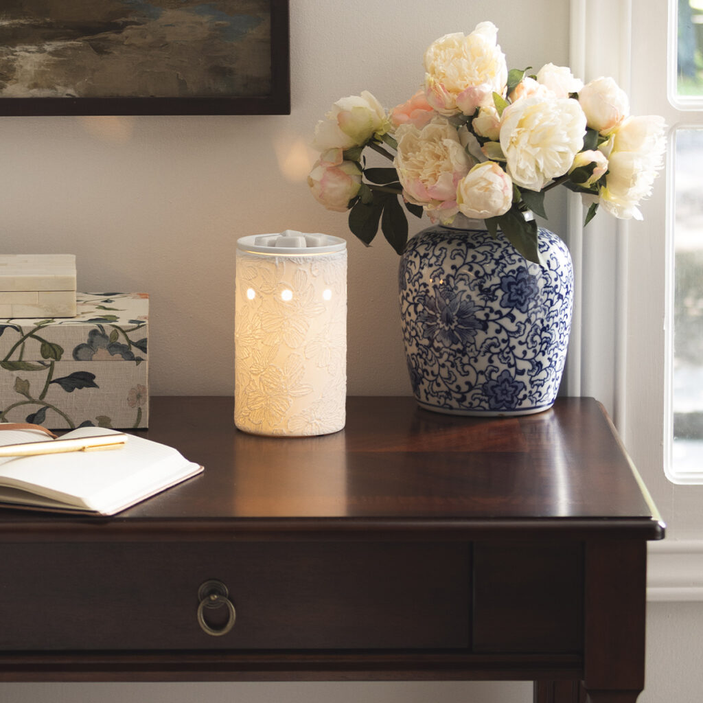 White Scentsy Lace & Light warmer glowing softly on a bedside table next to a floral vase.
