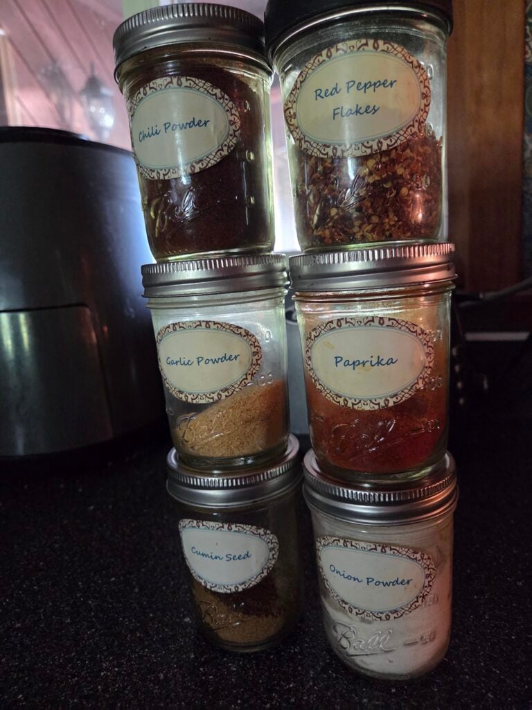 Homemade taco seasoning ingredients in labeled glass jars including chili powder, paprika, garlic powder, onion powder, cumin seed and red pepper flakes.