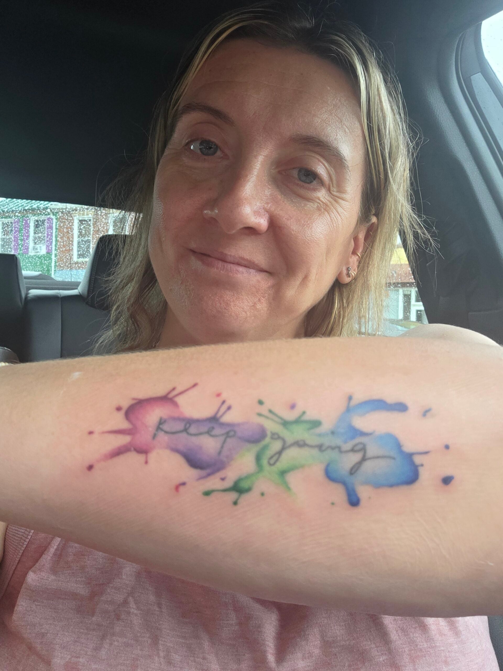 Amber Grillot showing her "Keep Going" watercolor tattoo with colorful paint splash artwork on her forearm.
