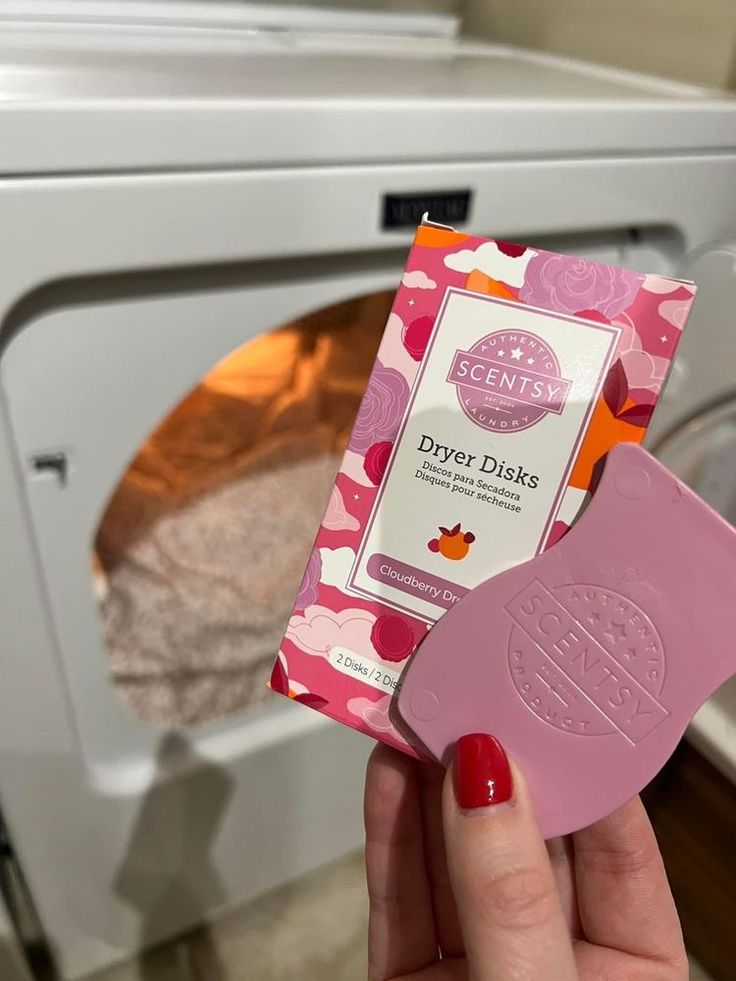 Hand holding a Scentsy Dryer Disk and Cloudberry Dreams Dryer Disks packaging in front of an open clothes dryer.