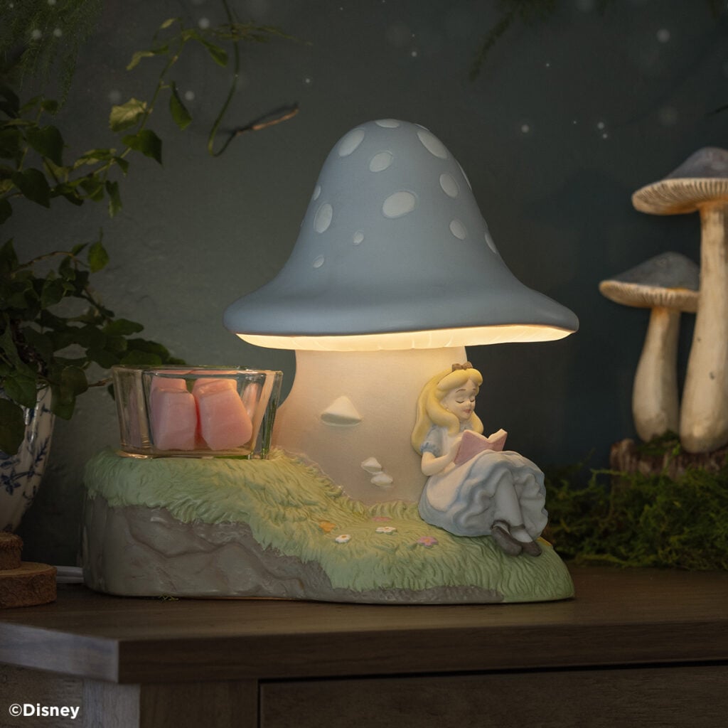Glowing Disney Alice in Wonderland Down the Rabbit Hole Scentsy Warmer, in the dark, shaped like a blue mushroom, with Alice reading beneath it and a glass dish holding pink wax cubes.