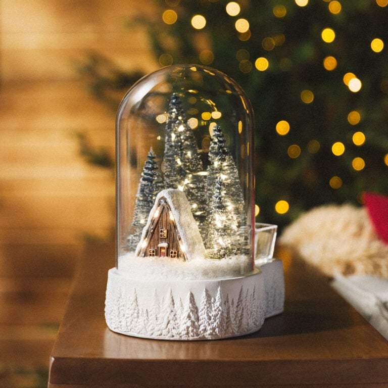 Magic of Winter Scentsy Warmer glowing in the evening, featuring a snow-covered cabin and lighted Christmas trees inside a glass dome.